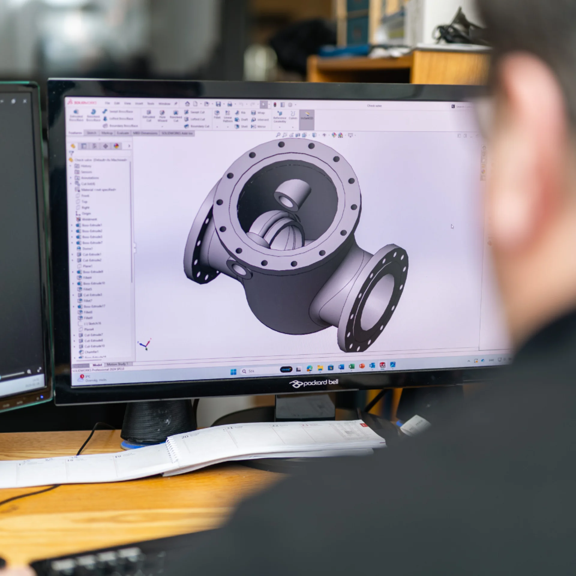 A person sits at a desk viewing a 3D model of a mechanical part on a computer screen, likely using CAD software, with papers and office items nearby.