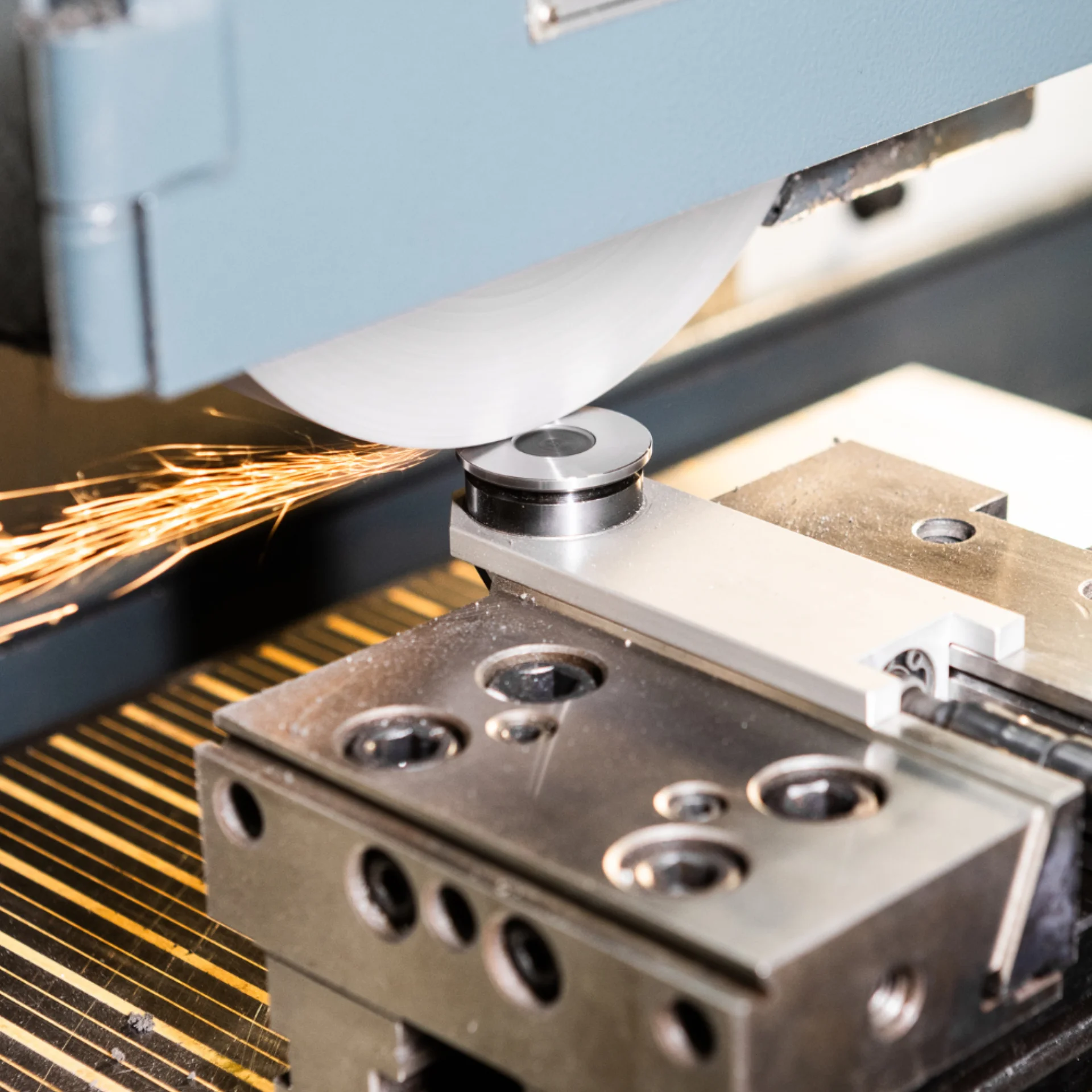 A grinding machine with a rotating wheel sharpens a metal component, producing bright sparks. The metal piece is secured in place by a clamp beneath the grinding wheel.