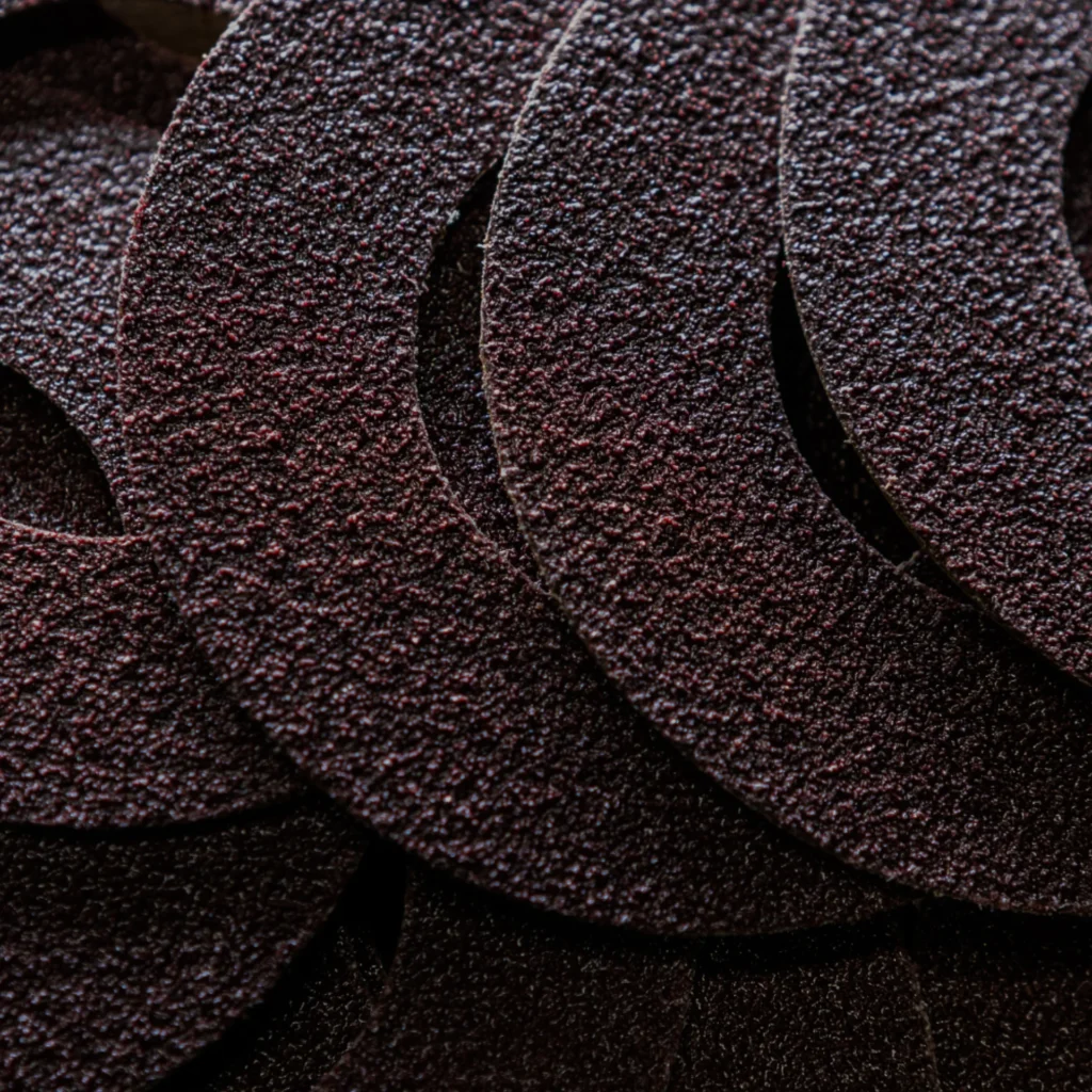 Close-up of overlapping circular pieces of coarse, dark brown sandpaper, showing their rough, gritty texture in detail.