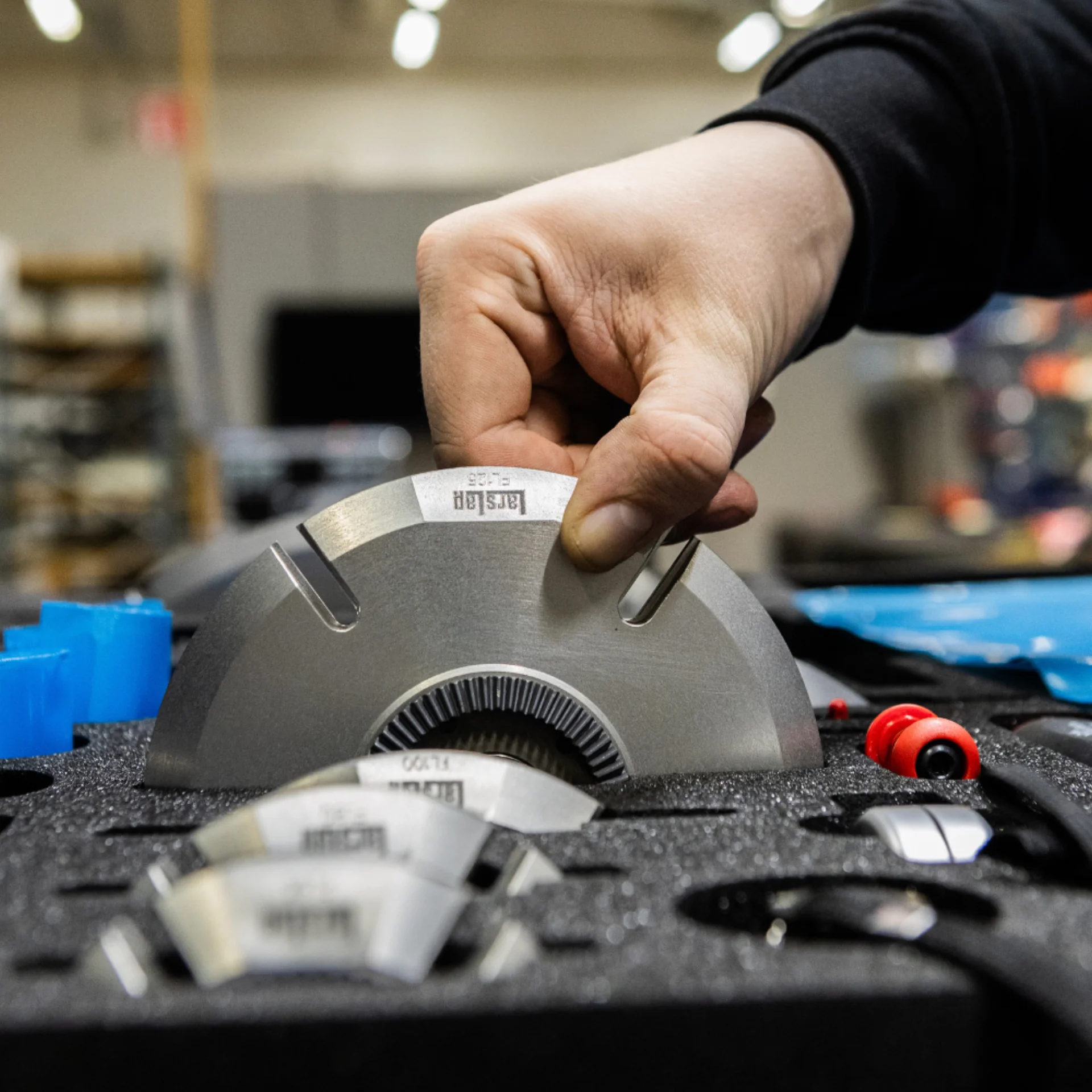 A hand lifts a large, semicircular metal blade from a foam case filled with similar precision tools, inside an industrial or workshop setting.